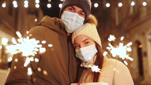 Beautiful couple in protective medical masks standing on the street holding sparklers in their hands of Christmas holidays. Romantic couple burning the sparklers at festive street fair 100fps — Stock Video