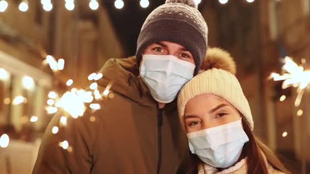 Couple in protective medical masks standing on the street holding sparklers in their hands of Christmas holidays. Romantic couple burning the sparklers at festive street fair. Happy Couple Outdoors — Stock Video