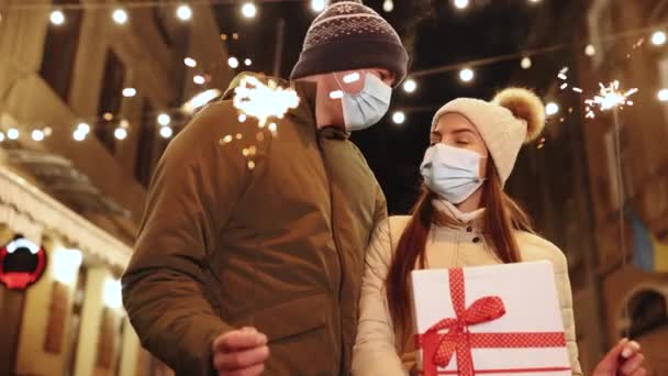 Pasangan dalam pelindung topeng wajah medis memegang dan melambaikan kembang api dengan merayakan Natal malam bersama-sama di dekorasi karangan bunga cerah. Pertukaran hadiah Natal. Perayaan Natal bersama — Stok Video