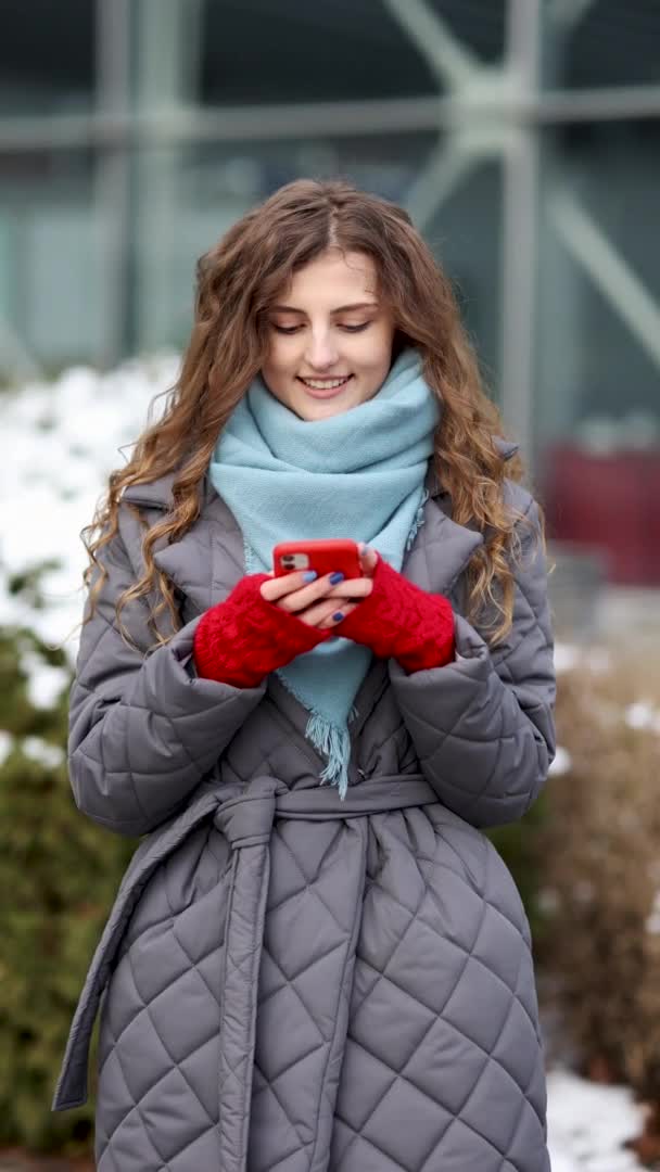 Vertikální záběr mladé kudrnaté ženy, jak stojí venku a píše sms na smartphonu. Usmívání je šťastný pocit. Zimní koncept. Veselé ženské smskování na smartphone venkovní. — Stock video