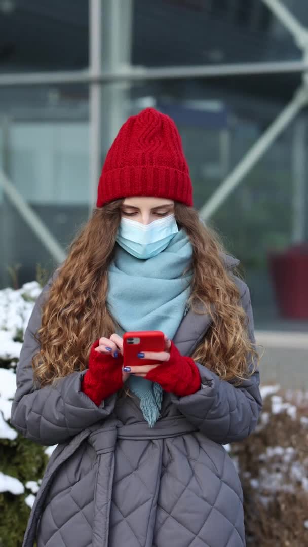 Vertikální snímek portrétu veselé kavkazské krásné kudrnaté ženy v masce, která si prohlížela smartphone, když stála na ulici. Radostná mladá žena píše na mobil. Orientace svislé obrazovky 4k. — Stock video