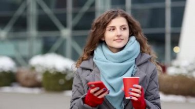 Kahve fincanı taşıyan mutlu, kıvırcık bir kadının portresi cep telefonundan mesaj atıyor. Sokak şehrine göz gezdiriyorum. 4k Ağır çekim.
