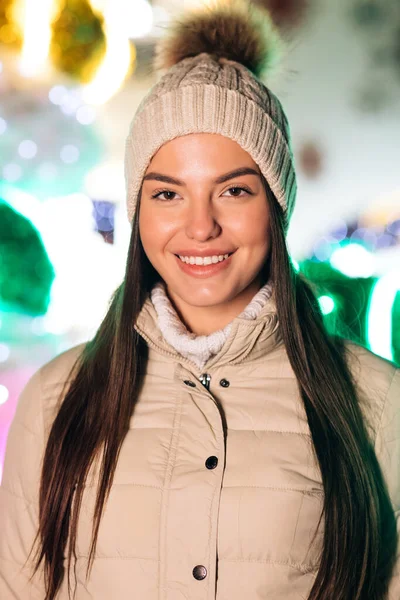Portret van vrolijke blanke vrouw met hoed die naar de camera kijkt en glimlacht rond kerstverlichting in het centrum van de stad. Positieve stemming vrolijke emotie buiten staan. Winter mooie straat gezicht buiten — Stockfoto