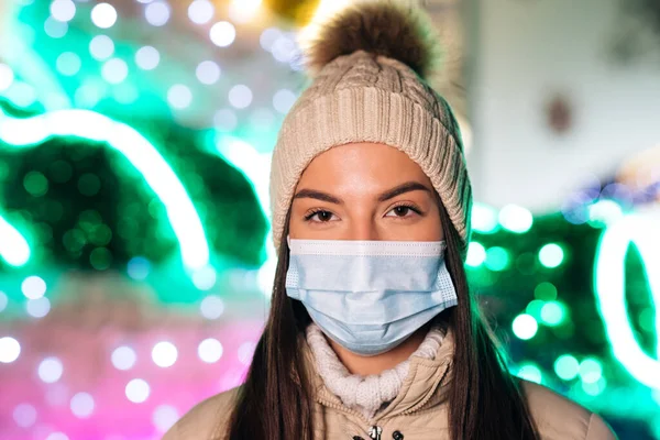 Potret perempuan muda Kaukasia cantik dengan topeng medis berdiri di kota Natal berdekorasi. Orang-orang sakit covid-19. Epidemic coronavirus. Virus flu korona pandemik. — Stok Foto