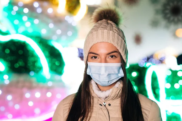 Epidemic coronavirus. Pandemic flu corona virus. Close up portrait of Caucasian young beautiful female in medical mask standing in decorated xmas city. Crowd people sick covid-19