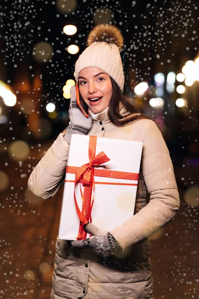 Verticaal Shot van portret mooie vrouw praten op de smartphone tijdens het staan op straat. Jonge vrouw met kerstcadeau aan de telefoon. Winterconcept. Verticale schermoriëntatie. — Stockfoto