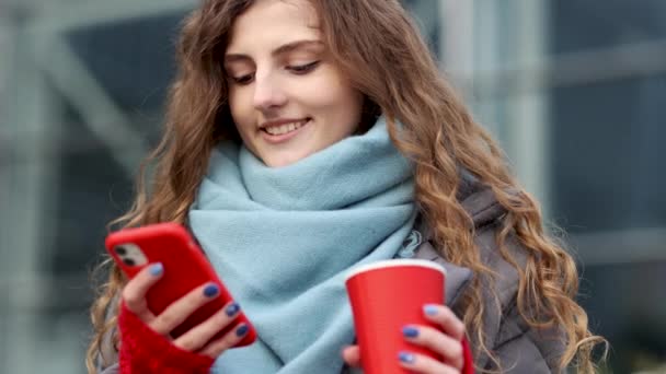 Kręcone włosy młoda dziewczyna śmieje się i czyta coś na jej smartfonie, trzymając filiżankę kawy w drugiej ręce. Kobieta spacerująca po mieście ulicy. Sieci społecznościowe — Wideo stockowe