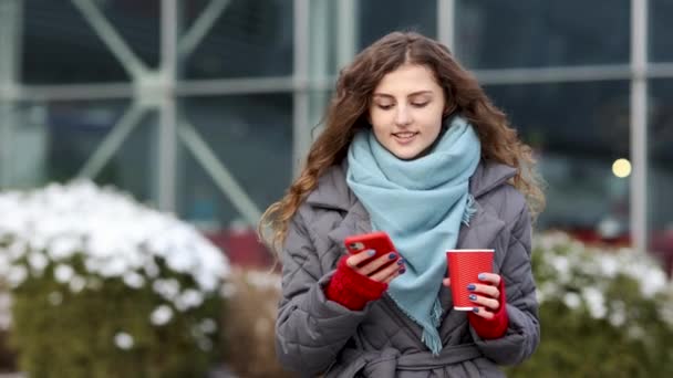 커피를 마시는 현대적 인 곱슬머리의 여성은 스마트폰을 사용 한다. 여성 초상화. 거리의 도시를 둘러 본다. 온라인으로, 소셜 네트워크로 — 비디오