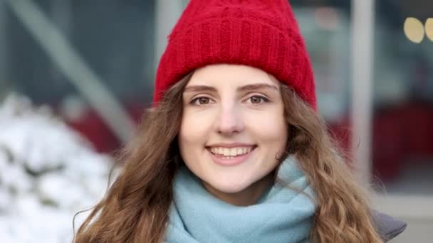 도시 중심가의 카메라를 보는 빨간 모자를 쓴 매력적 인 곱슬곱슬 한 미소를 짓는 젊은 여성의 모습이 행복하다. 완충 소녀. 길 위에 서. 귀여운 얼굴. 기운을 내라 — 비디오