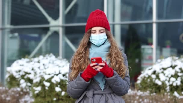 건강용 마스크를 착용 한 젊은 곱슬머리의 여성이 거리를 걸어 다닐 때 전화 텍스트 두루마리를 사용하여 인터넷 뉴스를 검색하고 19 코로나 바이러스 보호 도시를 검색 한다. — 비디오