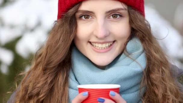 카메라를 보고 있는 예쁘고 곱슬머리의 우아 한 젊은 여성의 근접 사진. 여성 초상화. 손에 커피를 들고 빨간 모자를 쓴 소녀가 겨울에 거리에 서 있다. — 비디오