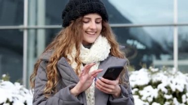 Kıvırcık kadın dışarıda akıllı telefon kullanıyor. Cep telefonunda yazan gülümseyen mutlu bir çalışan. Kış konsepti Neşeli, genç bir bayan. Yazıyor ve dışarıda akıllı telefondan kayıyor.