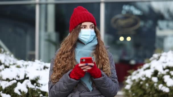 Portret wesołej kaukaskiej pięknej kręconej kobiety w masce przeglądającej się na smartfonie stojąc na ulicy. Radosna młoda kobieta pisząca na komórkę. Kręcone samice stojące na zimowej ulicy. — Wideo stockowe