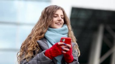 Dışarıda akıllı telefon kullanan kıvırcık saçlı kadın. Cep telefonunda yazan gülümseyen mutlu bir çalışan. Kış konsepti Neşeli, genç bir bayan. Yazıyor ve dışarıda akıllı telefondan kayıyor.