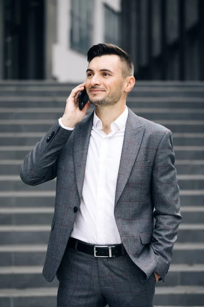 European businessman talking on mobile phone near office building background. Man is in dialogue, smiling, holding coffee cup on summer day. Vertical Screen. — Stock Photo, Image