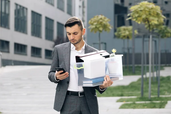 Sad young businessman uses phone texting scrolling tapping near office building. Male office worker in despair lost job. Look serious technology communication