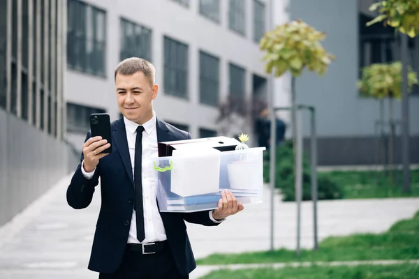 Young businessman with box of personal stuff uses phone texting scrolling tapping. Happy businessman stand smiling use phone near business center. Portrait suit career male office handsome technology