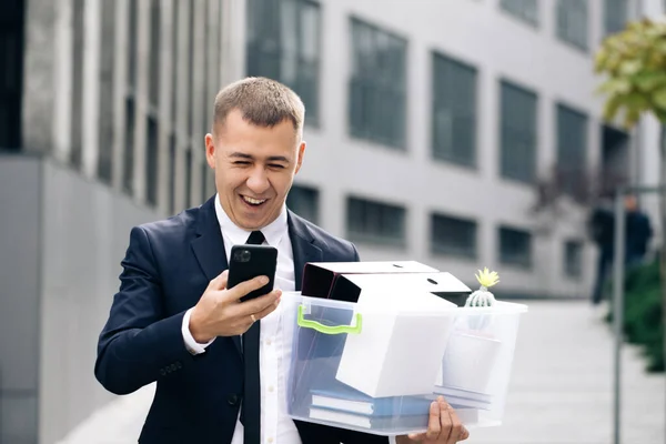 Businessman with box of personal stuff uses phone texting scrolling tapping. Happy businessman stand smiling use phone near business center. Portrait suit career male office handsome technology