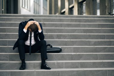 Elleri kafaya koymaktan yoruldum. Çaresizlik içindeki erkek ofis çalışanı işini kaybetti. Kötü ve rahatsız hissediyorum. Ofis binasının yanındaki merdivenlerde oturan üzgün genç işadamı.