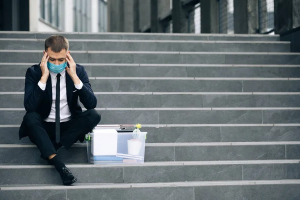 Sağlık maskeli, depresyondaki merdivenlerde bir kutu eşyayla oturan kovulmuş bir ofis çalışanı. Endişeli bir konsept. Umutsuzluk içindeki işsiz adam. İşsiz işadamı işini kaybetti