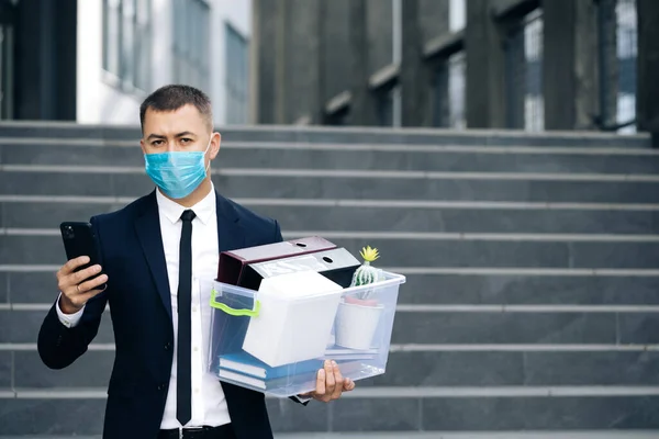 Businessman in medical mask with box of personal stuff walking the street use phone. Business style suit. Coronavirus outdoors social distancing. Finance and industry. Fired man lost job