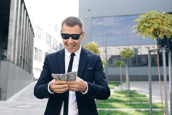 Homme riche en costume élégant portant des lunettes de soleil comptant l'argent et marchant près de l'immeuble de bureaux. Avoir l'air réussi, confiant — Photo