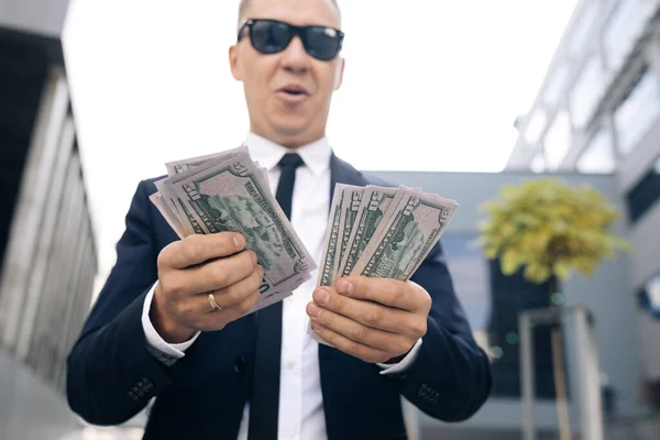 Un homme d'affaires prospère compte l'argent. Homme souriant profiter beaucoup près du bureau moderne. L'homme a gagné de l'argent facilement. Homme riche en costume élégant portant des lunettes de soleil comptant l'argent. — Photo