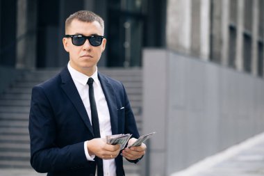 Başarılı Takım Elbiseli Genç İşadamı, Ofis Binası 'nın yanında duran Para Sayma Gözlüğü Giyiyor. Başarılı iş konsepti. İş hayatında mutlu ve yakışıklı bir adam dolar sayıyor.