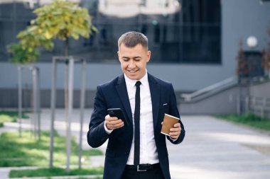 Modern Ofis 'ten çıkan şık bir iş adamı. Smartphone 'unu kullanıyor. Mobil Ekranda Kayıyor. Tasty Coffee içiyorum. Başarılı ve kendinden emin görünüyorsun. Yaz gününde kahve kupası tutmak