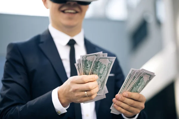 Een rijke man met een VR-headset die geld telt en lacht. Een jonge zakenman met een pak dollars. Rijkdom en succes concept. Nieuwe technologie biedt nieuwe 3D-dimensies. — Stockfoto