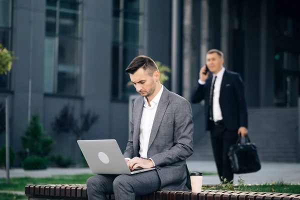 Hombre de traje trabajando con el ordenador portátil mientras está sentado en el banco. Concepto de trabajo remoto. Trabajo a distancia. Un hombre aislado con traje. Hombre de negocios escribiendo en el ordenador portátil al aire libre — Foto de Stock