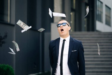 Dolarlar saçan mutlu yakışıklı adam. Para yağmuru, düşen dolarlar. Gözlüklü, takım elbiseli, zengin ve yakışıklı iş adamının portre resmi para saçıyor.