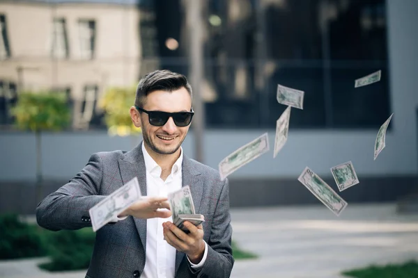 Succesvolle zakenman in een pak draagt een zonnebril Geld gooien in de camera staande in de straat in de buurt van Office Building. Geldregen, dalende dollars. Succesvol bedrijfsconcept — Stockfoto
