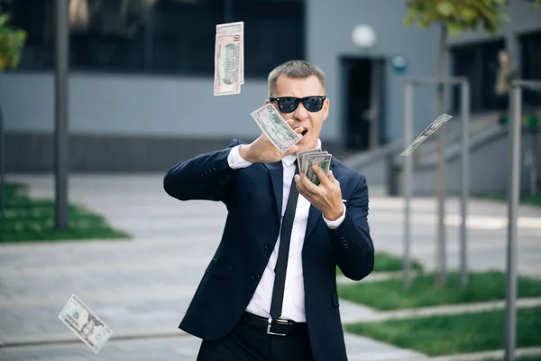 Para yağmuru, düşen dolarlar. Başarılı iş konsepti. Başarılı iş adamı takım elbiseli, güneş gözlüğü takıyor, kameraya para atıyor ofis binasının yanındaki sokakta duruyor.