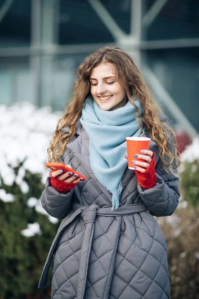 Vrouwelijk portret. Wandelen kijk rond op straat stad. Online zijn, sociale netwerken. Moderne stijlvolle krullende vrouw met kopje koffie, maakt gebruik van haar smartphone — Stockfoto