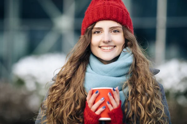 Πορτρέτο του όμορφου σγουρά καυκάσιος κομψό νεαρή γυναίκα κοιτάζοντας στην κάμερα. Γυναικεία πορτρέτα. Κορίτσι σε ένα κόκκινο ζεστό καπέλο με ένα φλιτζάνι καφέ στα χέρια της στέκεται στο δρόμο το χειμώνα. — Φωτογραφία Αρχείου