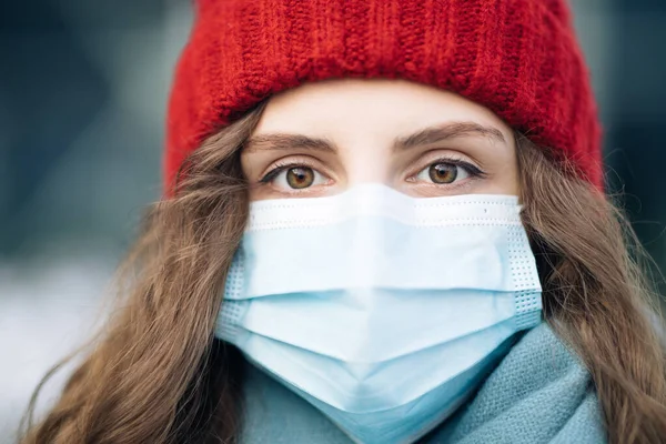 Portrait of Caucasian young curly female in medical face mask standing on winter street. Crowd people sick covid-19. Epidemic coronavirus. Pandemic flu corona virus