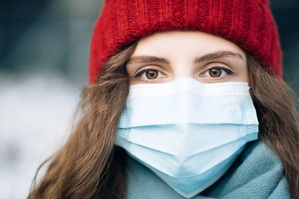 Close up of face calm serious curly haired woman wears protective mask to avoid contaminating coronavirus looks of city background. Health and safety concept. Curly female standing on winter street