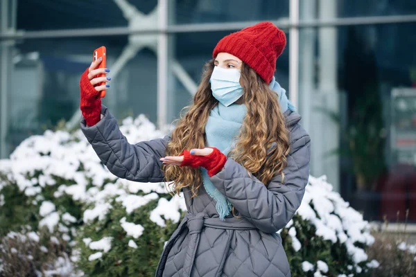 Vrouwelijke blogger in het gezicht masker is het maken van video praten voor haar sociale pagina op straat in de winter tijd. Een vrouw kijkt rond in de winter straat stad. Online zijn, sociale netwerken — Stockfoto