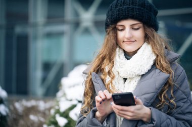 Dışarıda akıllı telefon kullanan bir kadın. Gülümseyen mutlu kız çalışan cep telefonunda daktilo ediyor. Neşeli genç bir bayan, dışarıda daktilo ediyor ve akıllı telefondan kayıyor. Kış konsepti