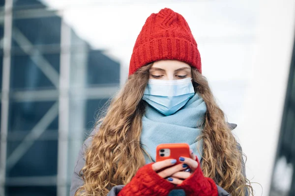 Een krullend vrouwtje op winterstraat. Blanke mooie krullende vrouw in masker browsen op de smartphone terwijl ze op straat. Joyful jonge vrouw sms 'en op mobiele telefoon — Stockfoto