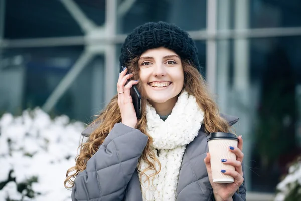 Σγουρά μαλλιά γυναίκα χρησιμοποιώντας το smartphone της και κρατώντας χάρτινο κύπελλο με καφέ. Νεαρή κοπέλα μιλάει στο κινητό της ενώ περπατάει στο δρόμο της πόλης — Φωτογραφία Αρχείου