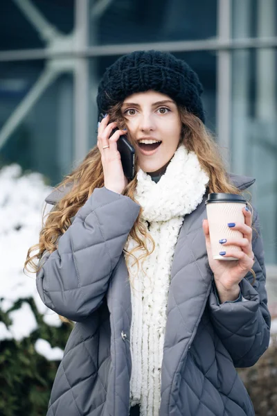 Νεαρή κοπέλα μιλάει στο κινητό της ενώ περπατάει στο δρόμο της πόλης. Γυναίκα χρησιμοποιεί το smartphone της και κρατώντας χάρτινο κύπελλο με καφέ — Φωτογραφία Αρχείου