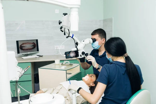 Dentista em luvas de látex examinando os dentes do paciente na clínica. Paciente deitado de boca aberta no consultório de dentistas. Estomatologista realizando exame usando um microscópio — Fotografia de Stock