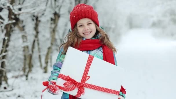 Niño en sombrero rojo con regalos de Navidad y regalos en la nieve. Diversión al aire libre de invierno. El niño juega en el parque nevado en Nochebuena. Feliz niña caucásica sonrisa y celebración de la caja de regalo. — Vídeo de stock