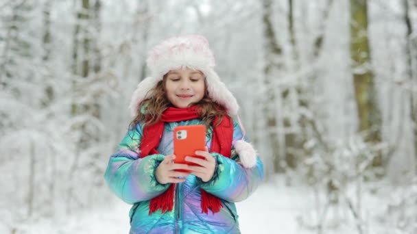 Κοριτσάκι τραβάει ένα αστείο βίντεο σε ένα smartphone. Ευτυχισμένο παιδί παίζει στο δάσος χειμώνα χρησιμοποιούν ένα smartphone ματιά στην οθόνη ενός κινητού τηλεφώνου, να παρακολουθήσετε κινούμενα σχέδια — Αρχείο Βίντεο