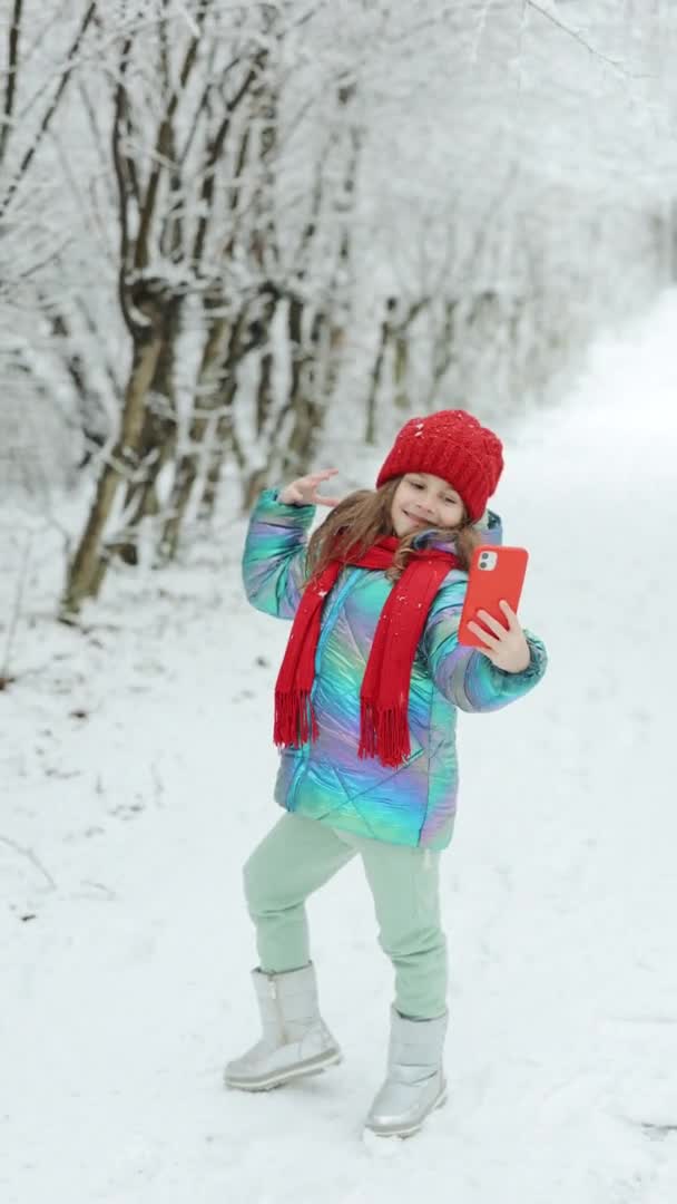 Küçük şirin bir kızın kış ormanında selfie çektiği dikey bir video. Çocuk konseptiyle kış seyahati. Sevimli, mutlu, gülümseyen bir kız çocuğu akıllı telefondan selfie çekmeyi sever. — Stok video