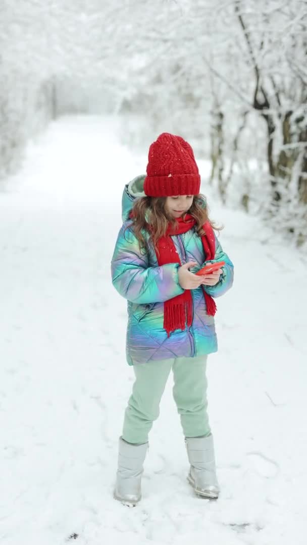 Tiro vertical de menina bonito miúdo usar telefone inteligente engraçado aplicativo máscara facial olhar para a tela celular sorri para algo engraçado. Lá fora. Os mortos do inverno, tempo frio, geada, neve — Vídeo de Stock