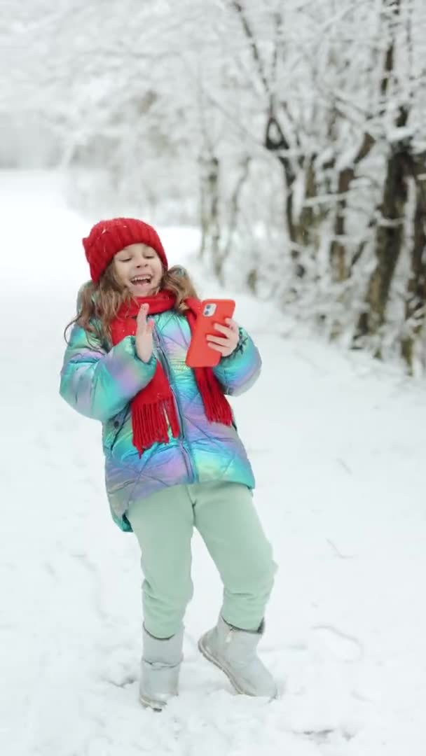 Pionowe wideo Happy cute little kid girl używać smartfona zabawna maska twarzy aplikacji spojrzeć na ekran komórkowy uśmiecha się na coś zabawnego. Na zewnątrz. Martwi zimy, zimna pogoda, mróz, śnieg. — Wideo stockowe