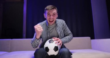 Yakışıklı beyaz adam endişeleniyor ve televizyonda futbol maçı için neşeleniyor. Gecenin geç saatlerinde kanepede oturuyor. Elinde top olan adama yaklaş ve spor kanalını izle..